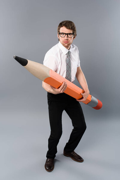 funny tense businessman in glasses holding heavy decorative pencil on grey background