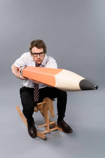 funny focused business man in glasses holding huge decorative pencil while riding rocking horse on grey background