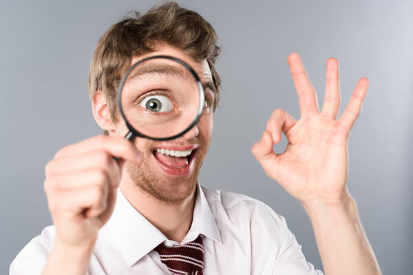 positive businessman with funny face expression looking in magnifier and showing ok sign on grey background