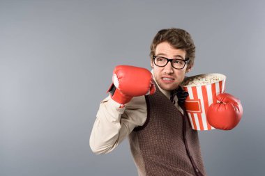 angry man in glasses and boxing gloves holding bucket of popcorn on grey background clipart
