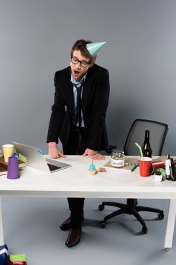 yawing businessman in party cap standing near workplace  clipart