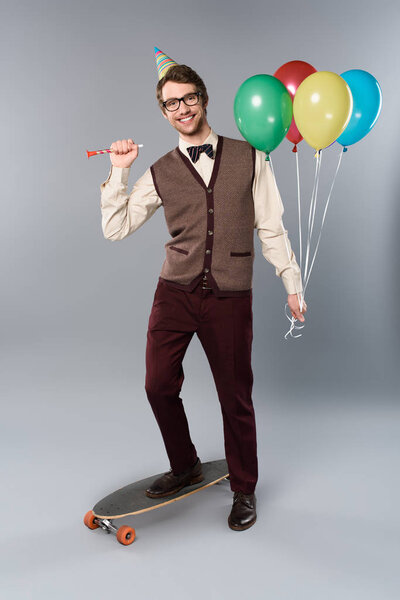 happy man in glasses and party cap holding multicolored balloons and party horn while standing on longboard on grey background