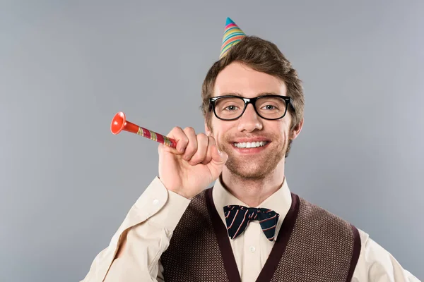 happy man in glasses and party cap holding party horn isolated on grey
