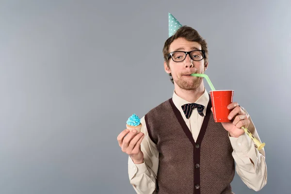 戴眼镜和党帽的人与搞笑的脸表情拿着纸杯蛋糕 喝从秸秆苏打水 — 图库照片