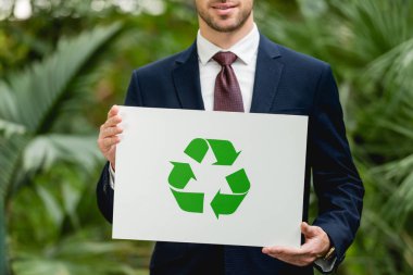 cropped view of smiling businessman in suit holding card with green recycling sign in greenhouse clipart