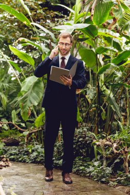 happy businessman in suit and glasses talking on smartphone and holding folder while walking in greenhouse  clipart