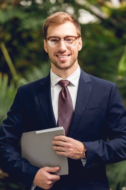 happy businessman in suit and glasses holding folder in greenhouse  clipart