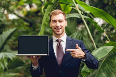 smiling businessman in suit and tie pointing with finger at laptop blank screen in orangery clipart