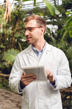 turunculuk dijital tablet kullanarak beyaz ceket ve gözlük gülümseyen yakışıklı bilim adamı