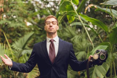 handsome businessman in suit and glasses with closed eyes holding rubber gas mask in orangery clipart