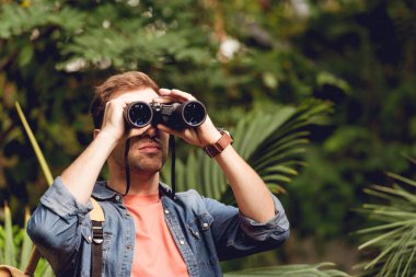 adult traveler looking through binoculars in tropical green forest clipart