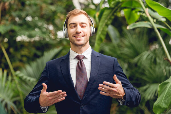 happy businessman in suit and wireless headphones breathing fresh air in greenhouse