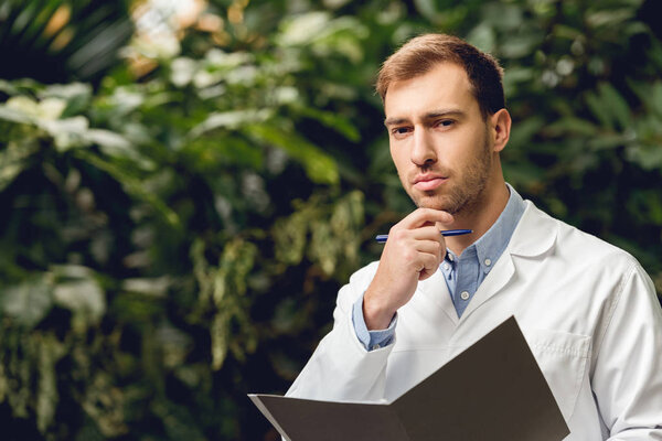 pensive scientist in white coat holding journal in green orangery