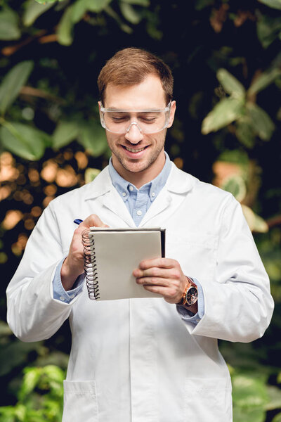 smiling scientist in white coat and goggles making notes in green orangery