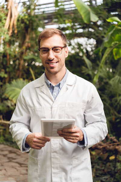 smiling handsome scientist in white coat and glasses holding digital tablet in orangery