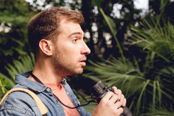 shocked handsome tourist with binoculars looking away in tropical forest