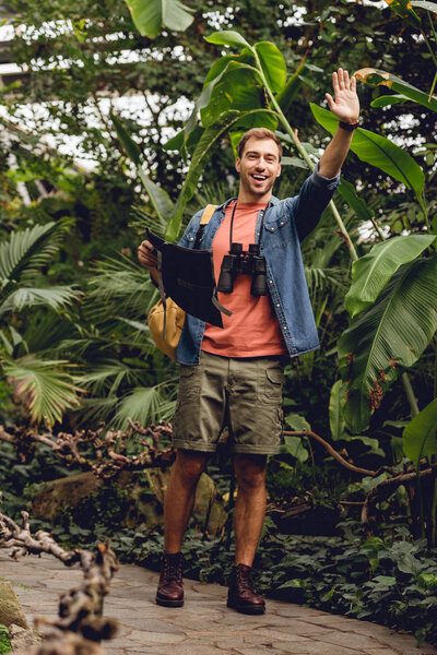 красивый счастливый человек с биноклем и рюкзаком, держащий карту и машущий рукой в зеленом лесу
