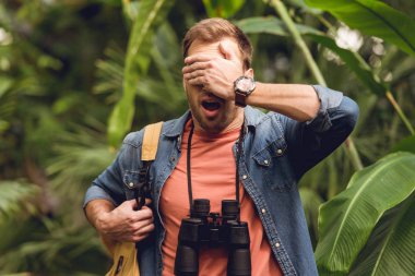 handsome scared traveler with binoculars and backpack putting hand on eyes in green tropical forest clipart