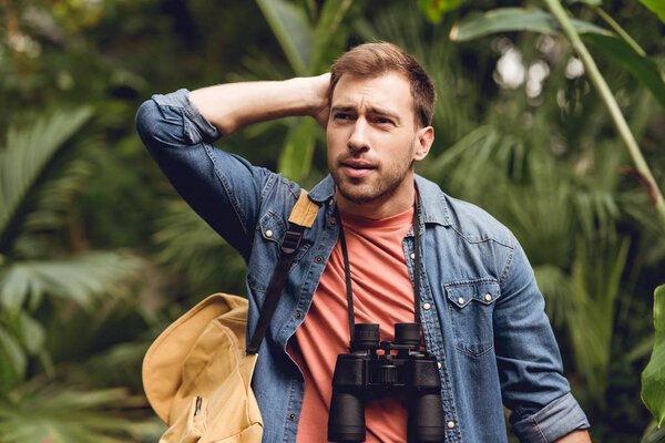 handsome tense traveler with binoculars and backpack in green tropical forest