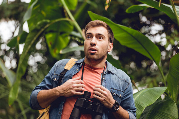 handsome scared traveler with binoculars and backpack in green tropical forest