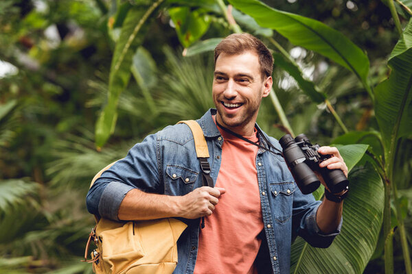 handsome smiling traveler with binoculars and backpack in green tropical forest
