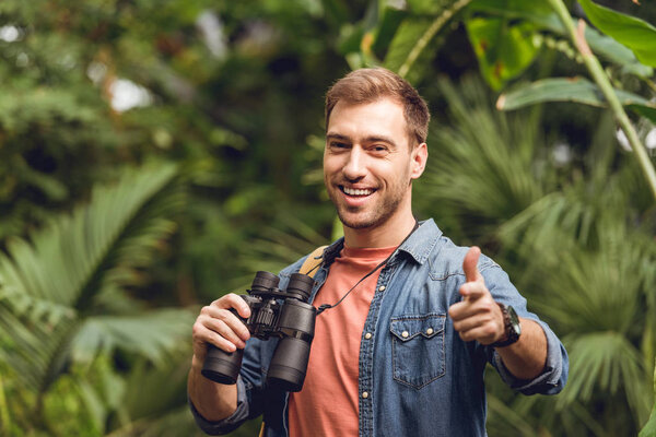 handsome smiling traveler with binoculars and backpack pointing with finger at camera in green tropical forest