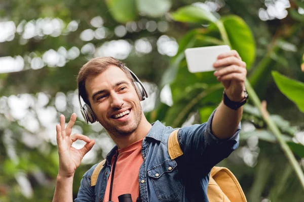 happy traveler in headphones taking selfie and showing okay sign in green tropical forest