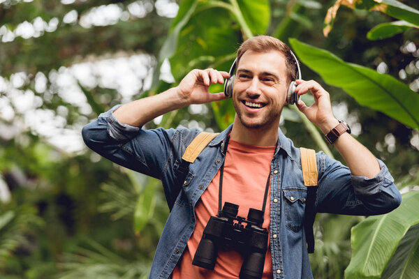 happy traveler with backpack and binoculars listening music in headphones in tropical forest