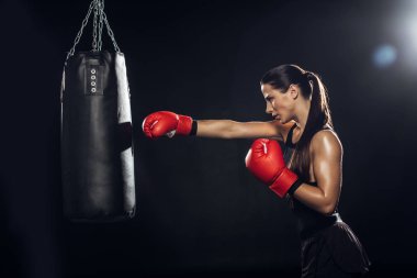 Side view of female boxer in red boxing gloves training with punching bag clipart