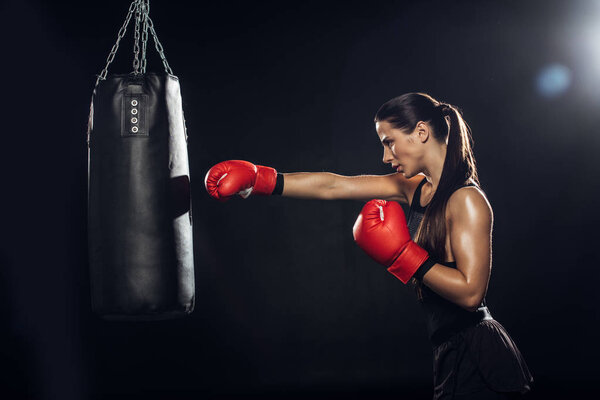 Side view of female boxer in red boxing gloves training with punching bag