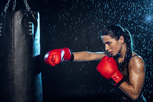 Pensive boxer in red boxing gloves training under water drops on black