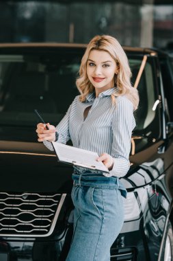 happy blonde car dealer holding clipboard and pen in car showroom  clipart