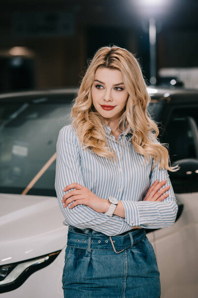 attractive blonde girl standing with crossed arms near automobile 