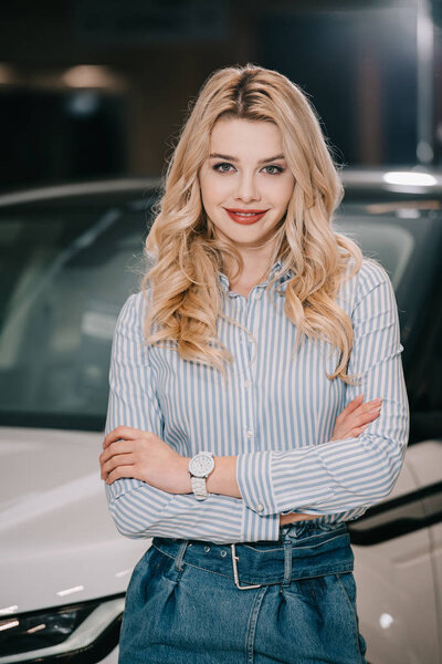 happy blonde girl standing with crossed arms near automobile 