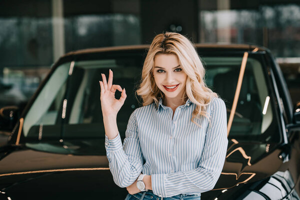 cheerful blonde woman showing ok sign near black automobile in car showroom  