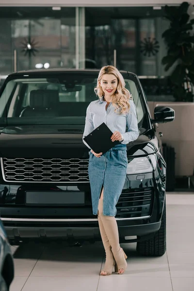 happy blonde car dealer holding clipboard while standing in car showroom