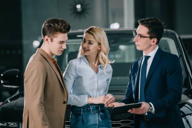 blonde woman looking at handsome man standing near car dealer with clipboard  clipart