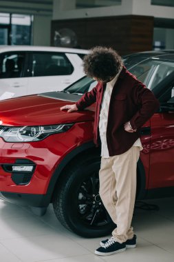 curly man looking at red automobile while standing in car showroom  clipart