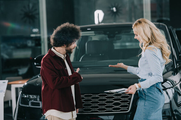 blonde car dealer showing black automobile to curly man 