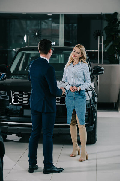 back view of car dealer holding clipboard with contract near attractive blonde woman 