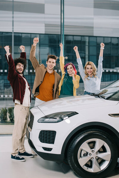 cheerful friends standing with hands above head near white car 