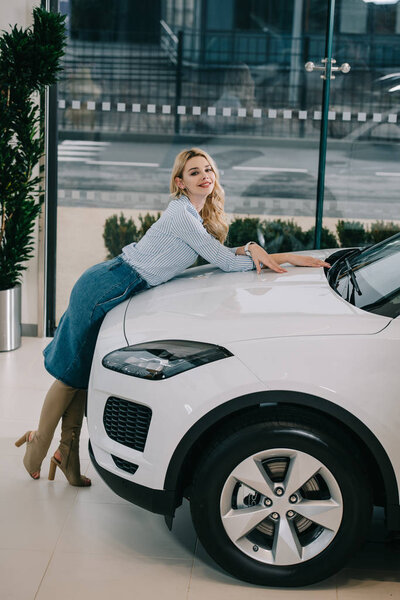 cheerful blonde girl standing near white automobile in car showroom 