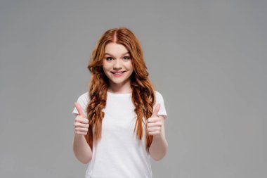beautiful redhead girl looking at camera, smiling and showing thumbs up isolated on grey with copy space clipart