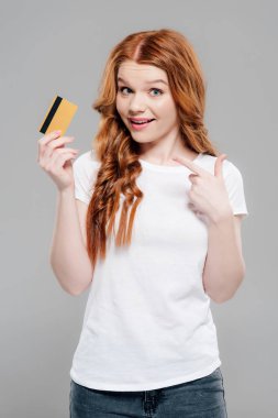 Güzel Redhead kız kamera bakıyor ve gri üzerinde izole kredi kartı parmak ile işaret