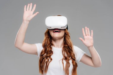 beautiful redhead girl in vr headset experiencing virtual reality and gesturing with hands isolated on grey clipart
