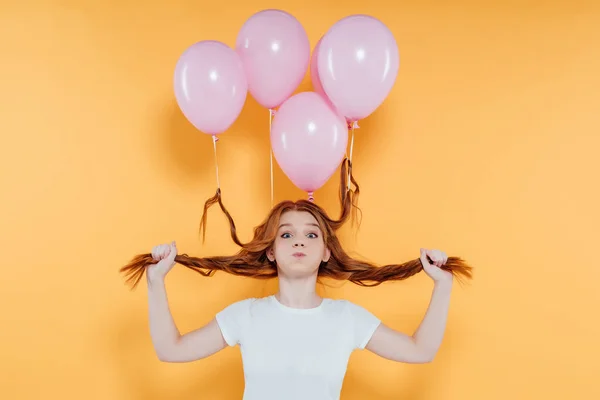 redhead girl with balloons tied to hair blowing cheeks and looking at camera isolated on yellow