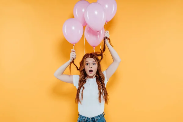 surprised redhead girl with balloons tied to hair looking at camera and posing isolated on yellow