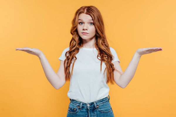 beautiful redhead girl looking at camera and showing shrug gesture isolated on yellow