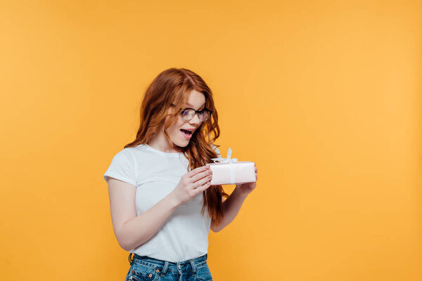 beautiful surprised redhead girl holding gift box isolated on yellow with copy space