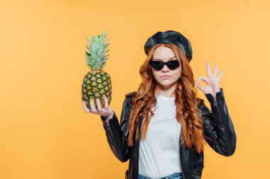 ciddi Kızıl saçlı kız deri ceket tutan ananas ve gösteren ok işareti izole sarı